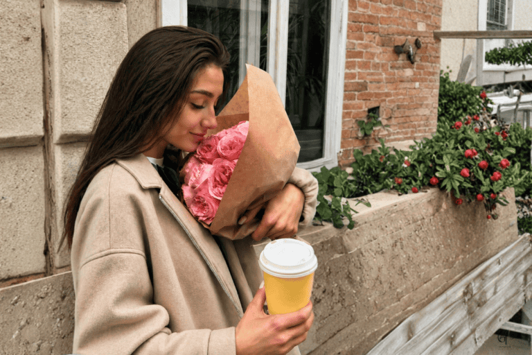 A person standing outdoors holding a bouquet of pink roses wrapped in brown paper and a yellow takeaway coffee cup, with green plants and red flowers in the background near a brick and concrete building.