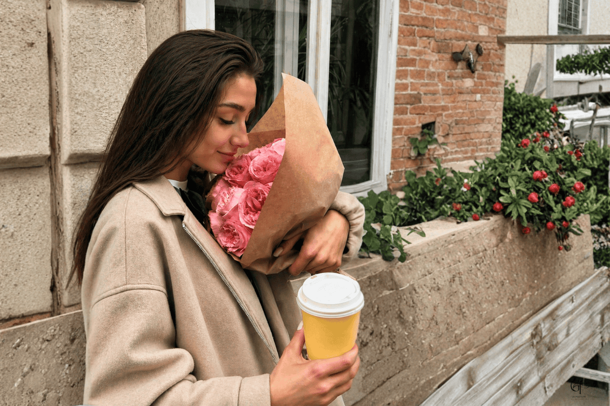 A person standing outdoors holding a bouquet of pink roses wrapped in brown paper and a yellow takeaway coffee cup, with green plants and red flowers in the background near a brick and concrete building.