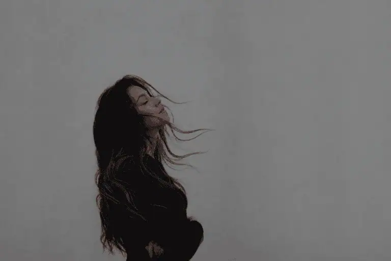 Moody minimalist portrait of a young woman with windblown dark hair standing against a soft grey background, symbolizing healing, anxiety recovery, and emotional resilience.