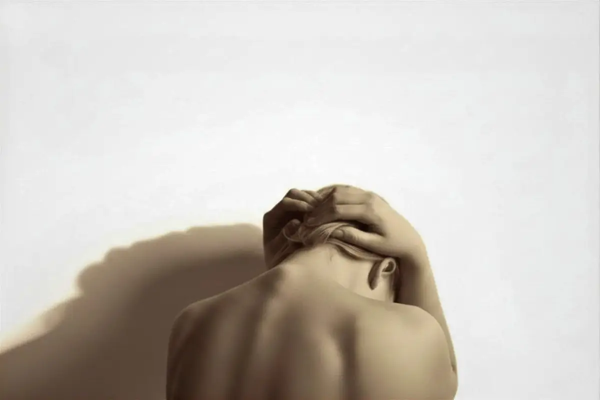 A person with light skin is shown from behind, head bowed forward, hands clasped over the top of the head. Their spine and shoulder blades are visible, and soft light creates gentle shadows on the plain background.