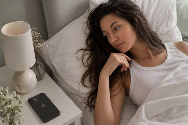 A woman resting peacefully in bed beside a gratitude journal and phone, symbolizing the choice between mindful morning rituals and phone scrolling.