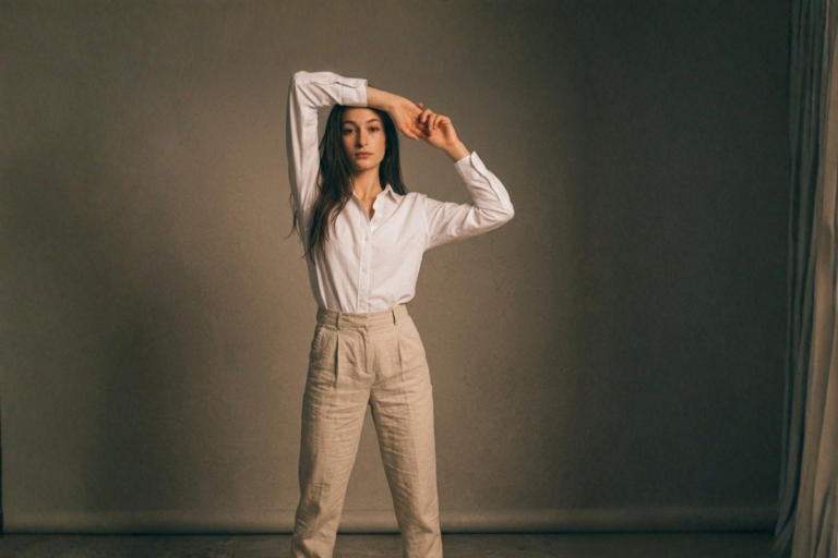 A person standing against a soft, neutral background, wearing a white button‑up shirt and high‑waisted beige trousers. Their arms are raised in a relaxed, open pose, creating a calm, minimalist, body‑aware atmosphere.