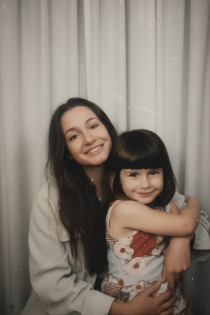 Portrait-style photo of an adult woman smiling while holding a young child on her lap. The child hugs the adult and wears a patterned dress, while the adult wears a light top. Soft indoor lighting and a neutral curtain backdrop create a warm, intimate family moment.