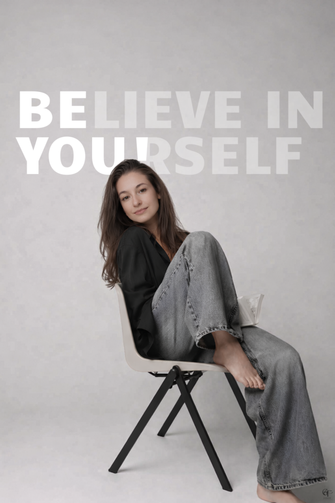 A person sitting sideways on a chair in relaxed posture, wearing casual clothing against a minimalist gray background.