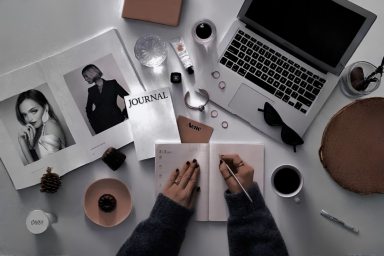 An overhead flat‑lay of a minimalist workspace with hands writing in a planner, surrounded by a laptop, sunglasses, coffee, water, rings, skincare items, a Chanel jar, a pine cone, and a magazine. The scene visually contrasts Energy Management and Time Management through a calm, intentional planning environment.