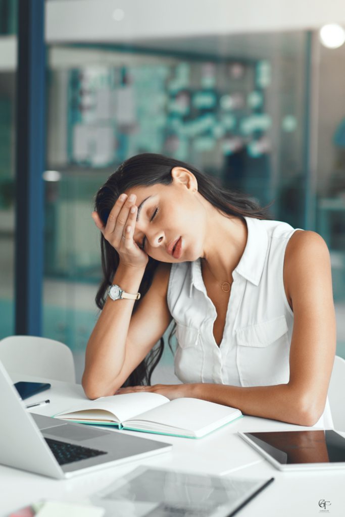 Exhausted woman resting her head in her hand at an office desk with an open journal and laptop