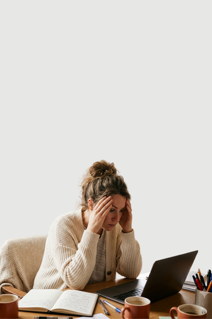 Woman sitting at a desk with her head in her hands, looking overwhelmed in front of an open laptop and journal