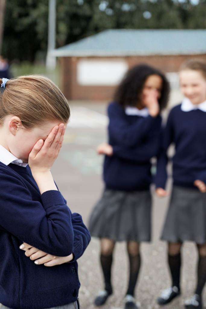 Schoolchildren standing outside, one child covering their face while others interact, representing subtle social exclusion and the hidden signs of bullying.